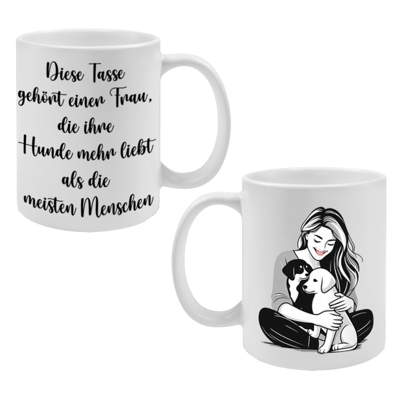 Weiße Kaffeetasse mit schwarzem Aufdruck: Auf einer Seite der Spruch „Diese Tasse gehört einer Frau, die ihre Hunde mehr liebt als die meisten Menschen“, auf der anderen eine schwarz-weiß-graue Illustration einer stilisierten Frau mit zwei Hunden, die sich liebevoll an sie schmiegen. Geschenkidee für Hundemamas